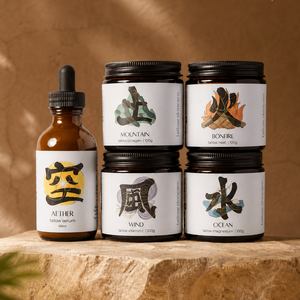 Four glass jars and one amber dropper bottle arranged on a stone slab against a warm brown backdrop. The products have white labels with black text and symbols, including Japanese characters and words like Mountain, Fire, Wind, and Water.