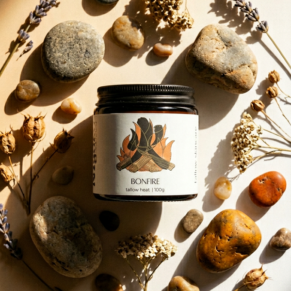 Jar of Bonfire tallow heat product surrounded by stones and dried plants on a light background