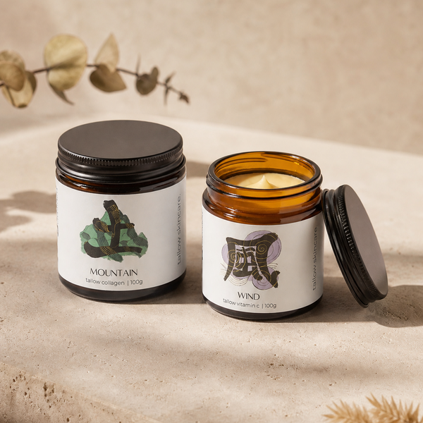 Two amber glass tallow jars with white labels and black lids are arranged on a beige stone-like surface. One jar is closed with a black lid, and the other is open, showing the candle wax and lid resting beside it.