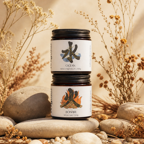 Two stacked glass tallow jars with black lids and white labels, placed on smooth stones among dried flowers and pebbles in a warm beige setting. The top label shows a dark blue wave-like symbol and the word OCEAN, while the bottom label shows a campfire graphic and the word BONFIRE.