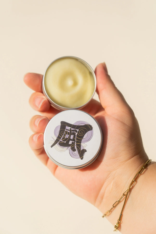 Hand holding a small round container of tallow cream with a visible brand logo on a beige background