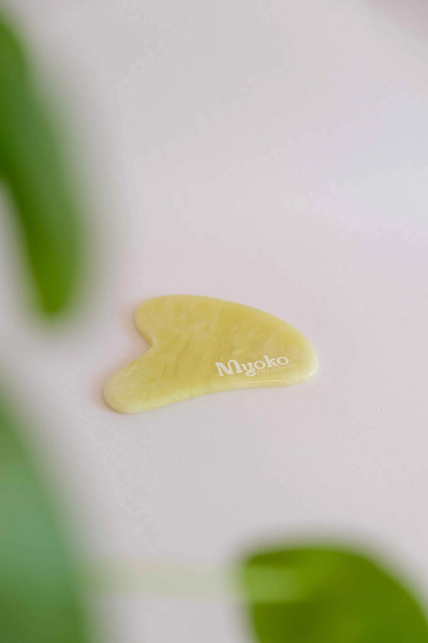 Yellow gua sha tool with 'Myoko' branding on a blurred green plant background