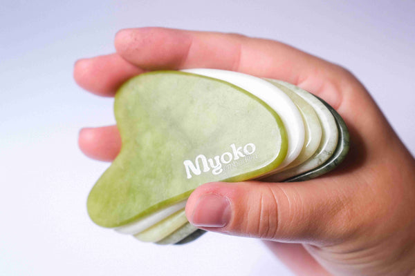 Hand holding a stack of green and white gua sha tools with 'Myoko' branding.
